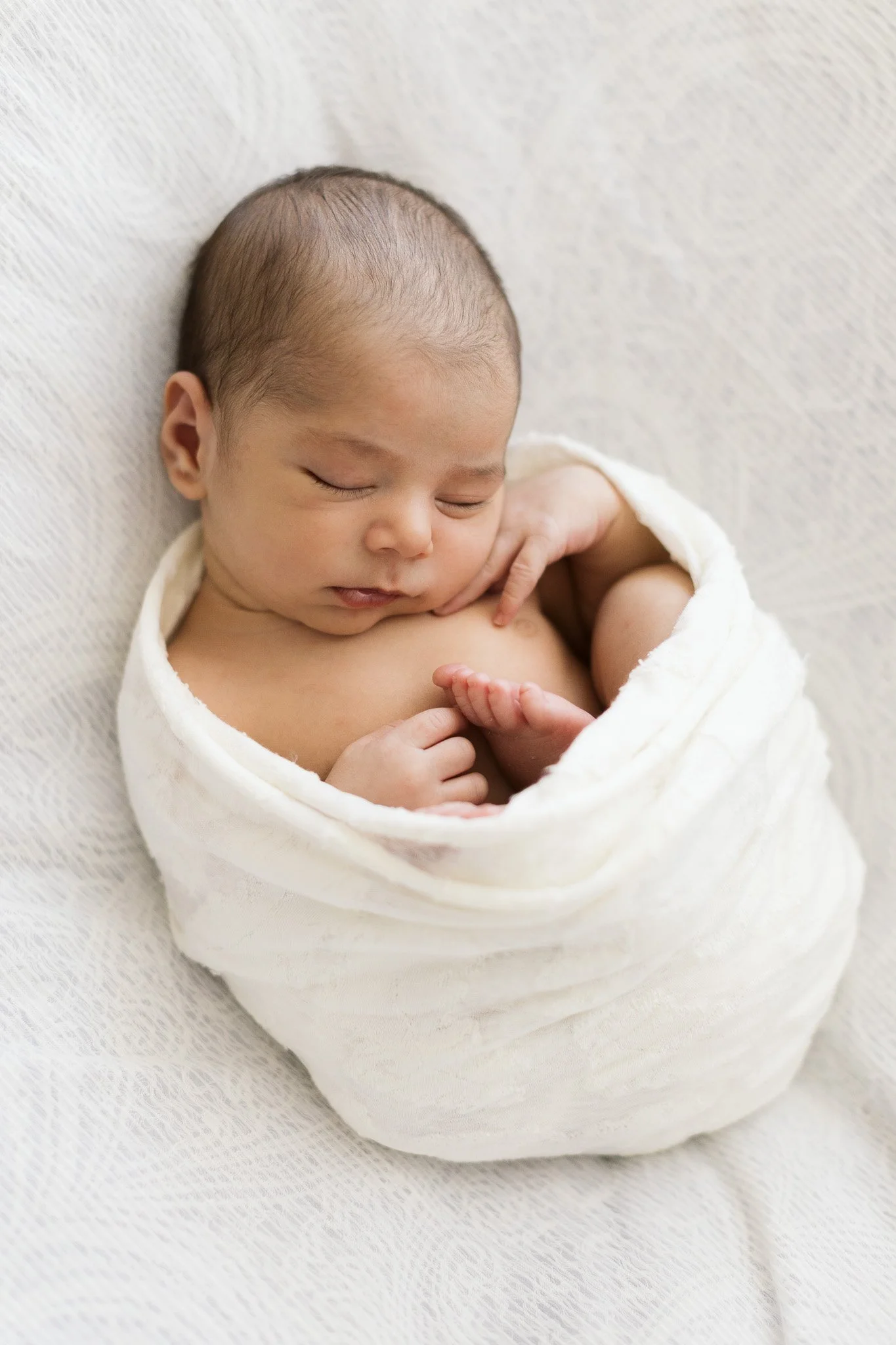 Neonato avvolto in una coperta bianca, dormiente, sullo sfondo di tessuto chiaro.