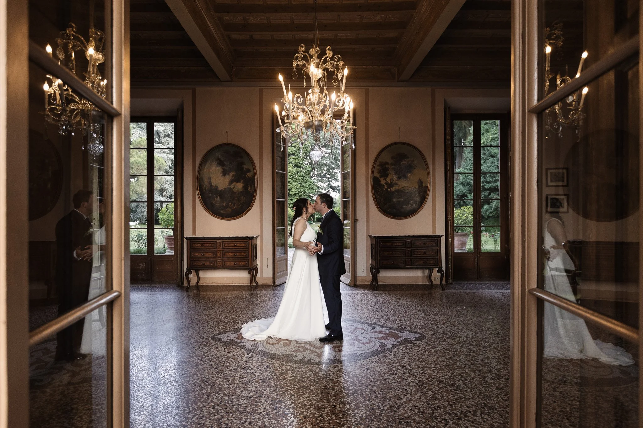 Coppia di sposi in abito da matrimonio che si baciano nel centro di una sala elegante con grande lampadario e pareti decorate con quadri ovali, visti attraverso due porte di vetro.