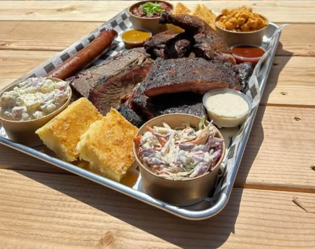 Barbecue platter with ribs, brisket, sausage, cornbread, potato salad, coleslaw, baked beans, and various dipping sauces on a metal tray outdoors.
