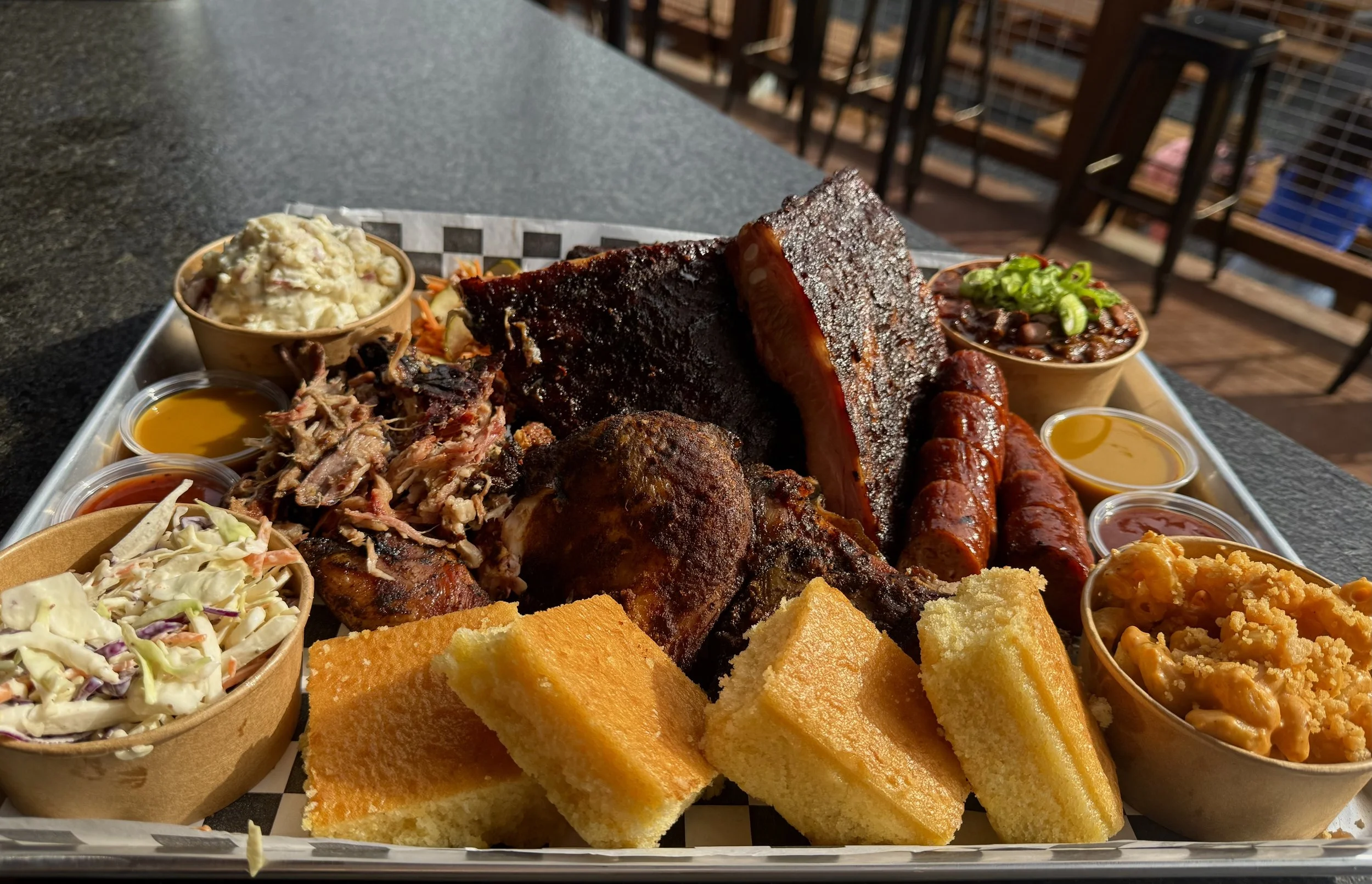 Barbecue platter with ribs, sausage links, fried chicken, macaroni and cheese, coleslaw, baked beans, potato salad, cornbread, and various barbecue sauces on a tray.