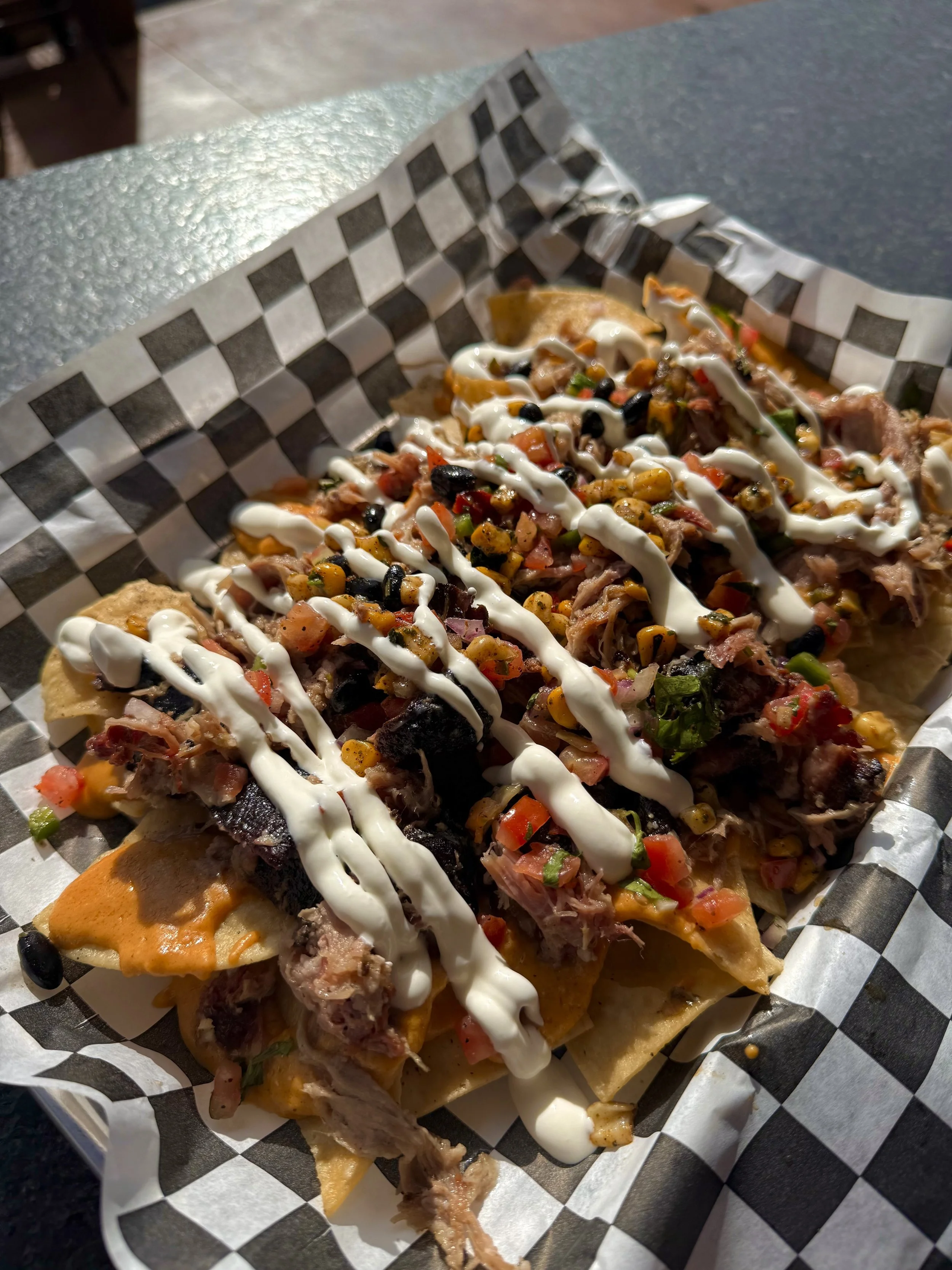 Close-up of loaded nachos with shredded meat, sour cream, and diced vegetables on black and white checked paper