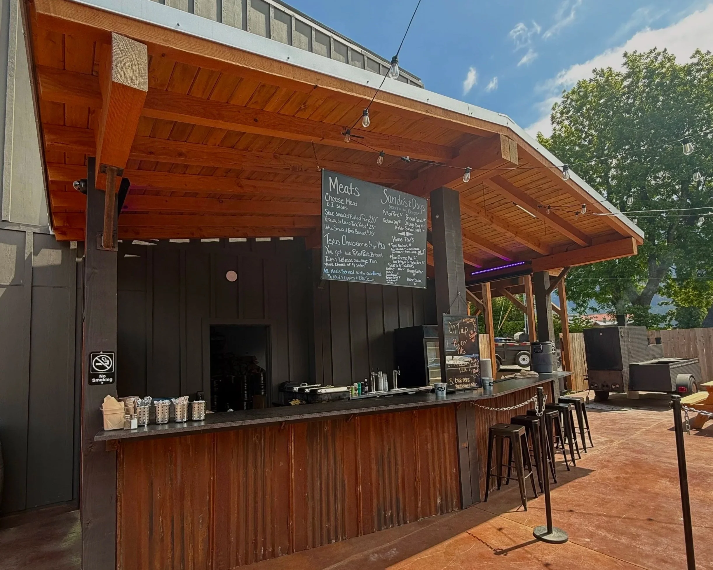 Outdoor food stand with a wooden roof, black walls, and a menu board listing meats, sandwiches, dogs, and sides, with bar stools in front.