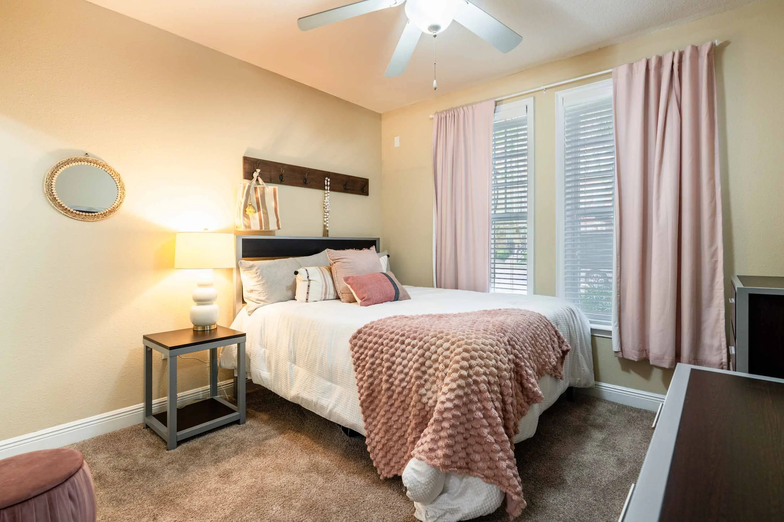 A cozy bedroom with a bed dressed in white linens, pink pillows, and a pink textured blanket. There is a nightstand with a white lamp beside the bed, and a mirror with a woven frame hanging on the wall. The room has two large windows with white blind