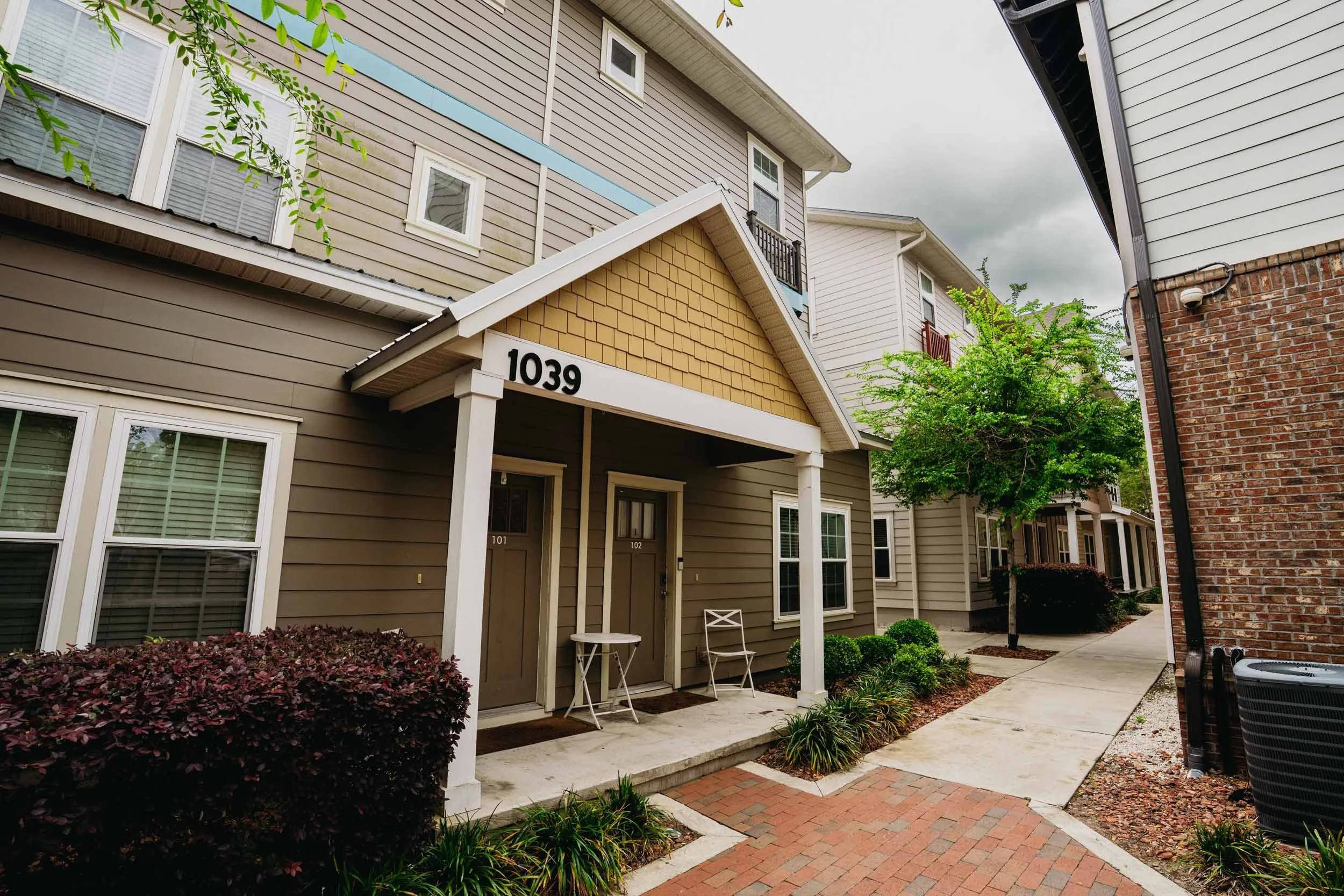 A multi-story residential building with beige and white siding, a small covered front porch, and a brick sidewalk in front. There are green bushes, small trees, and bushes along the pathway, with a cloudy sky overhead.