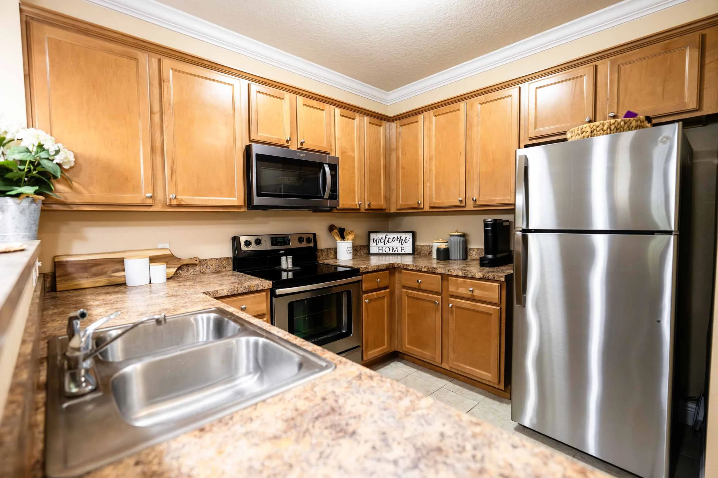 Kitchen with wooden cabinets, stainless steel refrigerator, microwave, and oven. Countertop with a sink, a potted flower, and decorative items.