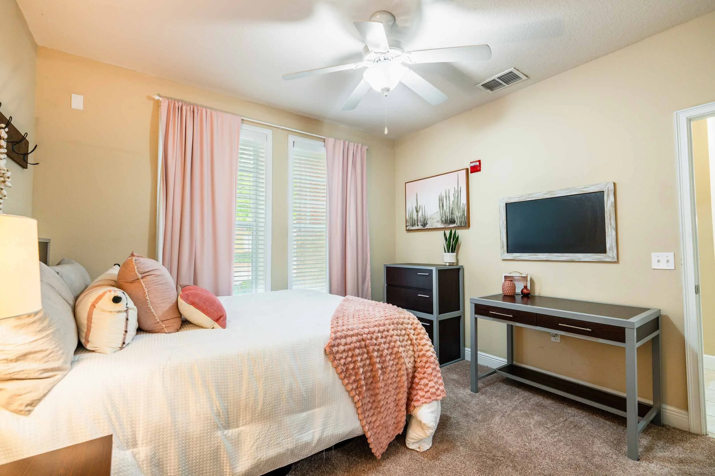 A bedroom with a bed on the left, decorated with pillows and a blanket. Two windows with pink curtains behind the bed. A ceiling fan with lights is on the ceiling. On the right, there's a dresser with a plant, framed desert cactus artwork, a blackboard, and a small table with decorative items. The room has beige walls and carpeted floor.