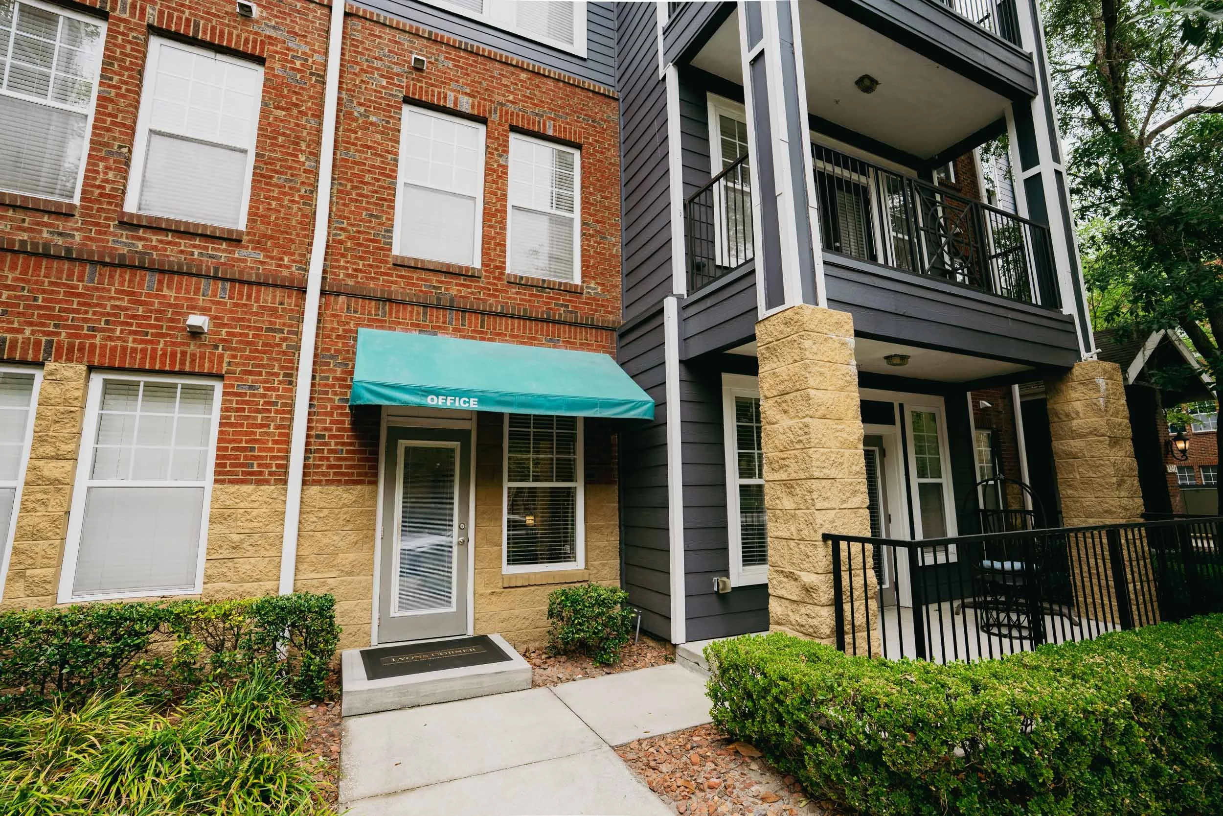 Exterior view of a multi-story residential building with a brick facade and a black wooden balcony. There is a green awning labeled 'OFFICE' over a door, and the entrance is surrounded by small bushes and a concrete walkway.
