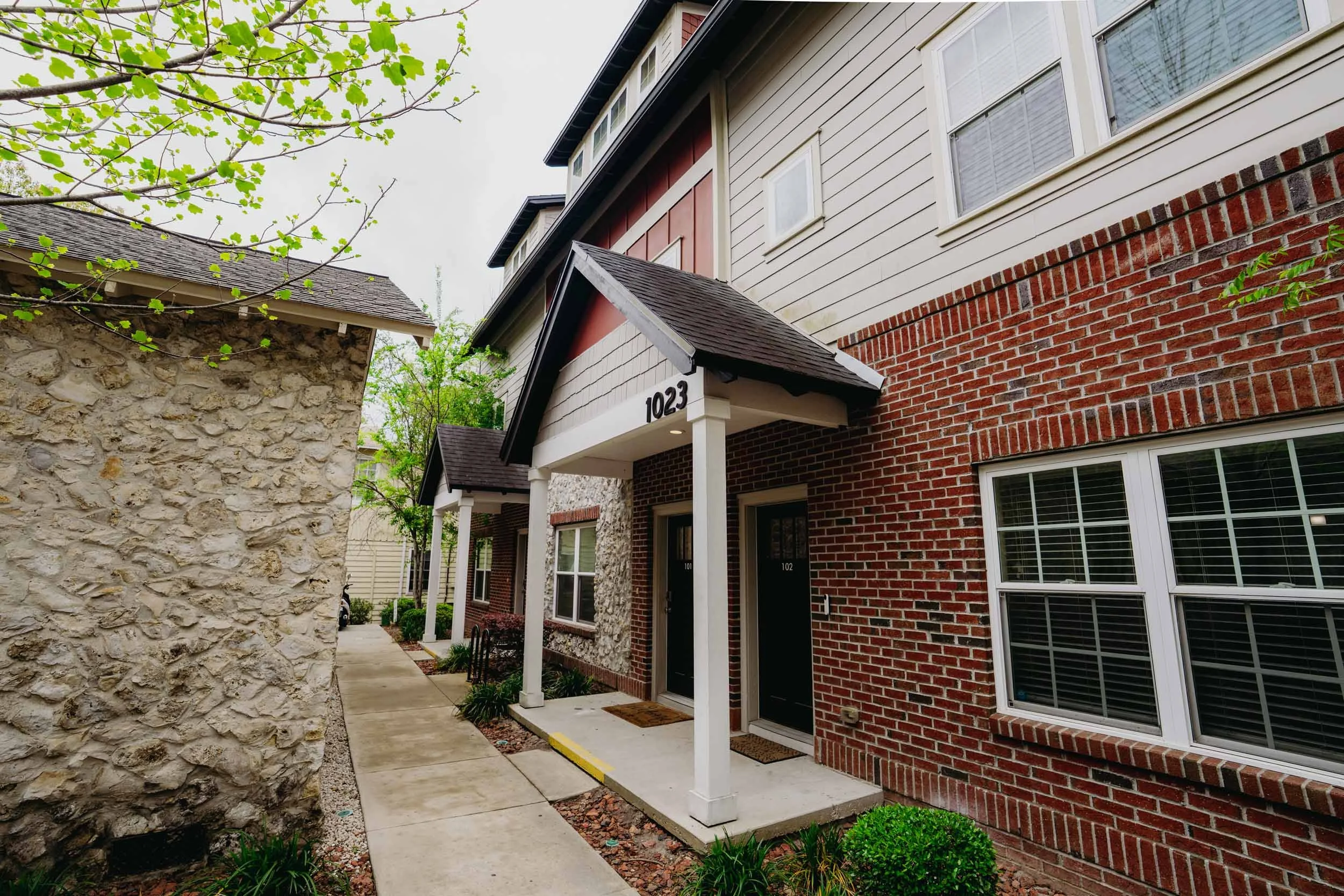 Exterior view of residential buildings with brick and siding facades, a sidewalk, and green plants, with a tree in the foreground.