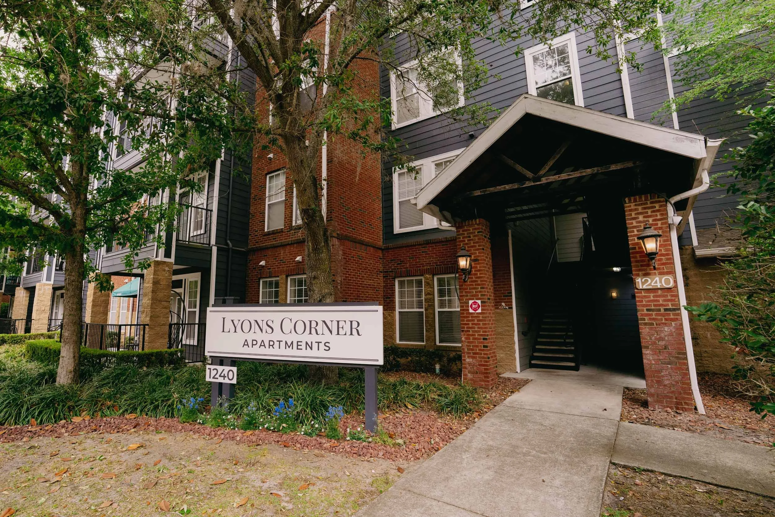 The image shows the entrance to Lyons Corner Apartments, a residential building with a brick and dark gray siding exterior. There is a white sign with black lettering near the sidewalk indicating the building's name and address, 1240. The entrance ha