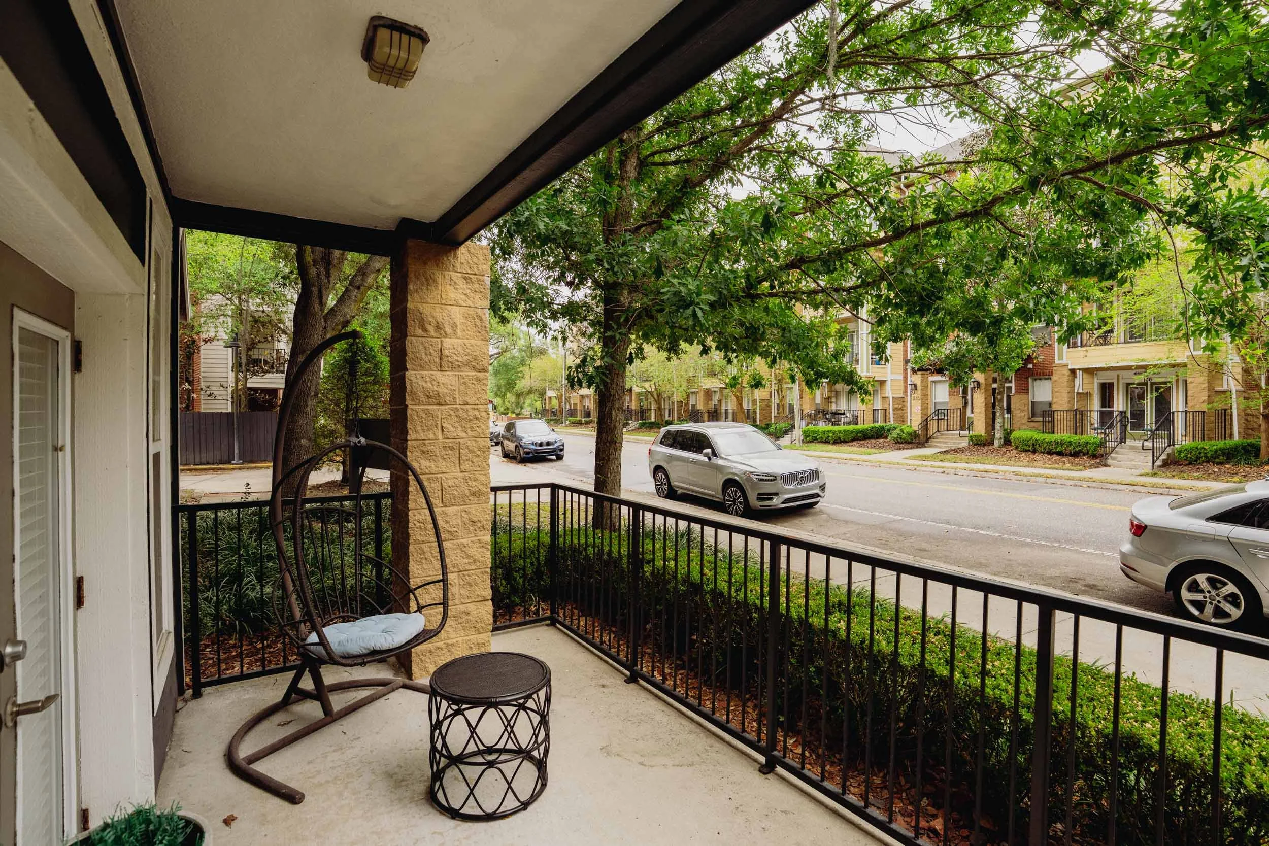 A covered front porch with two chairs and a small table, overlooking a street with parked cars and trees in a residential neighborhood.
