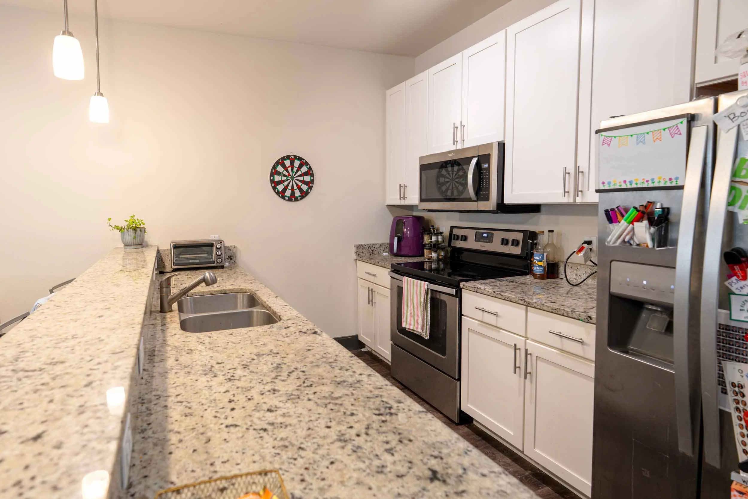 A kitchen with white cabinets, stainless steel appliances, and a speckled granite countertop. A dartboard hangs on the pale wall, and a potted plant is on the counter. The kitchen includes a microwave, toaster oven, purple coffee maker, and various small items. An overhead light with two pendant fixtures is visible.