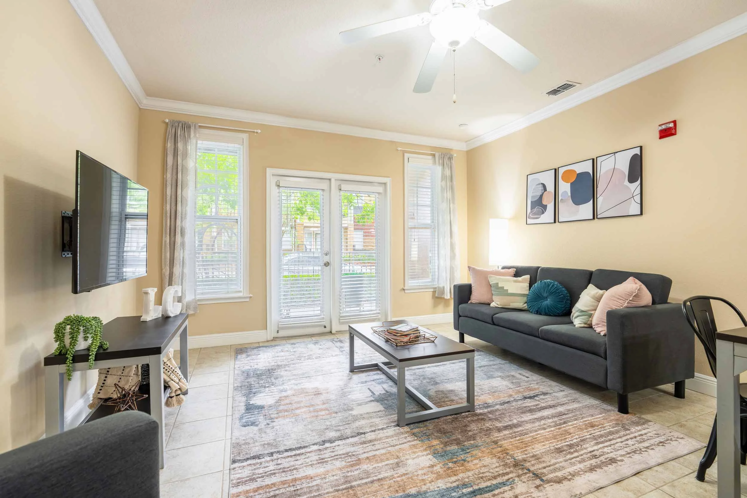 A living room with beige walls, a black sofa with colorful pillows, a rectangular coffee table, a wall-mounted flat-screen TV, and a doorway with glass doors leading outside. There are framed abstract art pieces on the wall, a floor lamp, and several