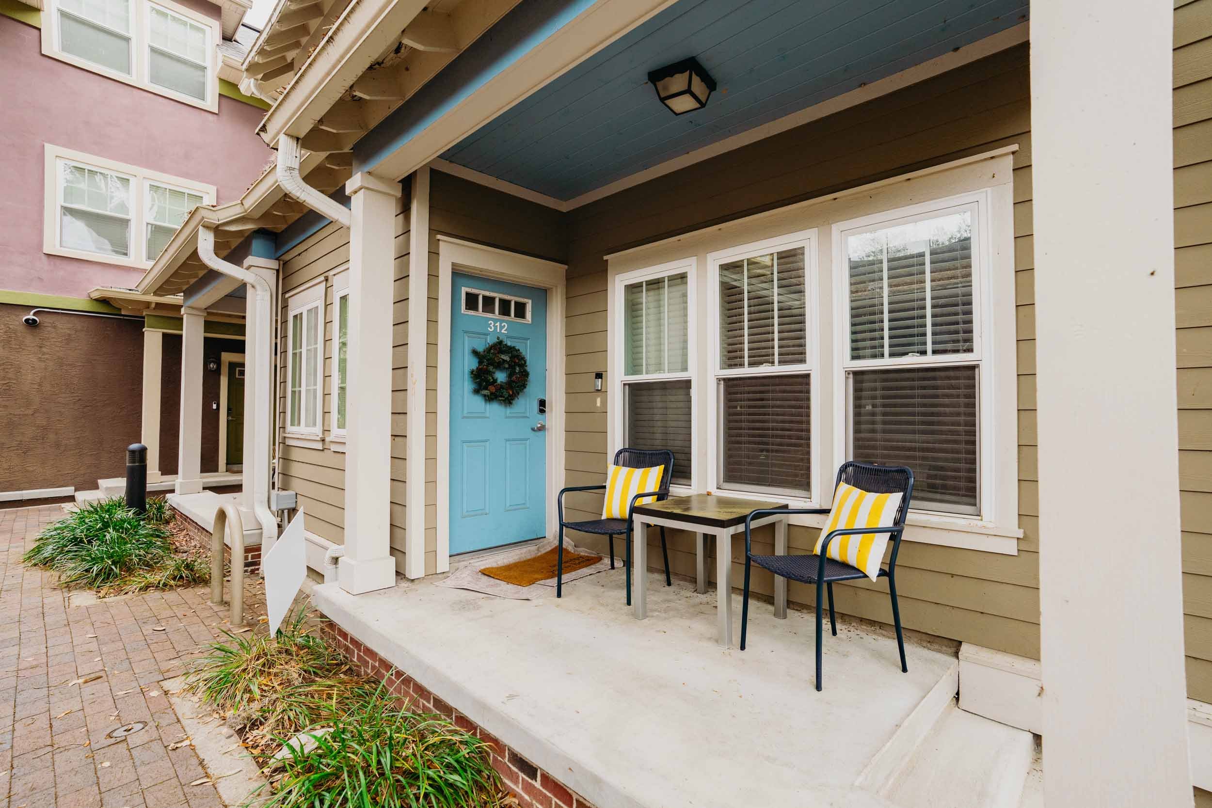 Front porch of a house with a light blue door with the number 312, a holiday wreath, two black chairs with yellow and white striped cushions, a small wooden table, large windows with blinds, and a covered area with a blue ceiling.