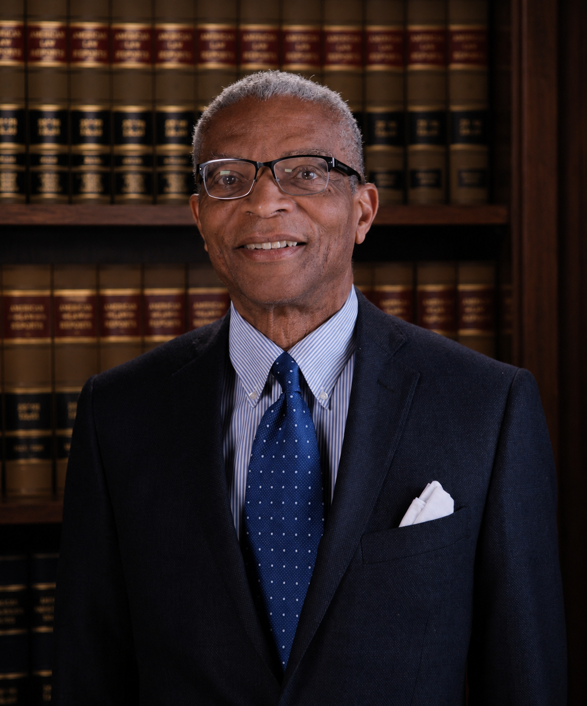 Portrait of an older African American man wearing glasses, a dark suit, a light striped shirt, a blue polka dot tie, and a white pocket square, smiling against a dark background.