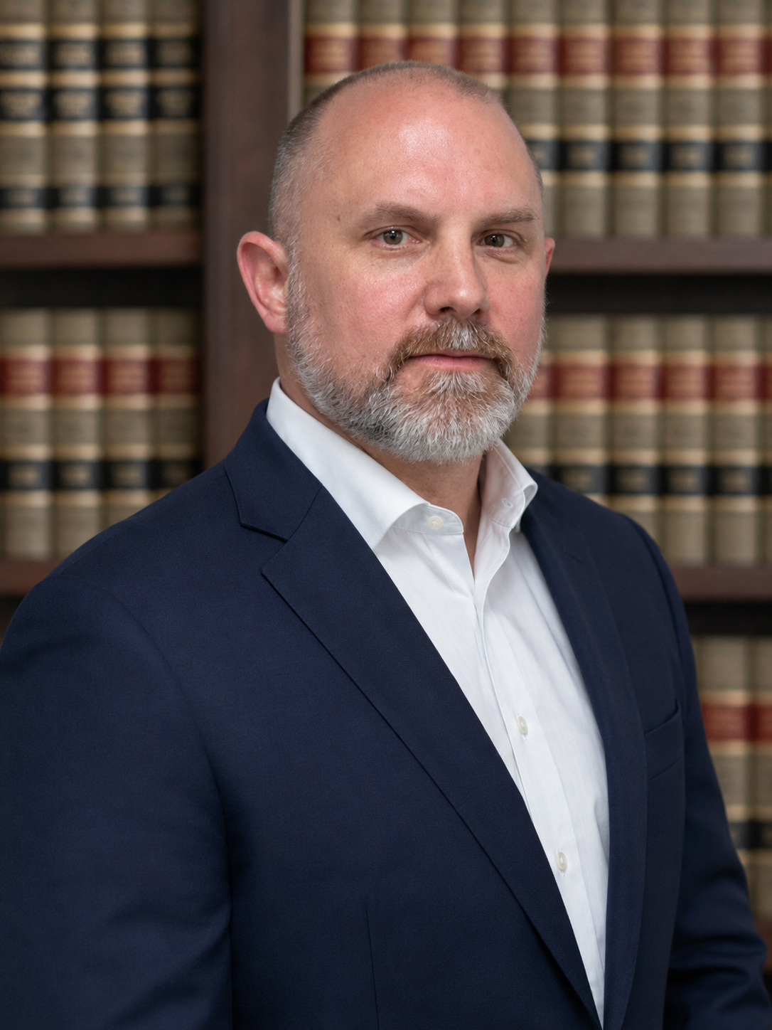 Professional headshot of a man with a bald head and gray beard, wearing a dark blazer and white shirt, against a light gray background.