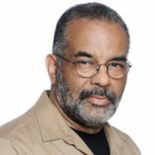 Close-up portrait of a middle-aged man with glasses, a beard, and short curly hair, wearing a beige jacket over a black shirt.