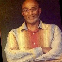 A man standing with his arms crossed, smiling, wearing glasses and a multicolored striped shirt.