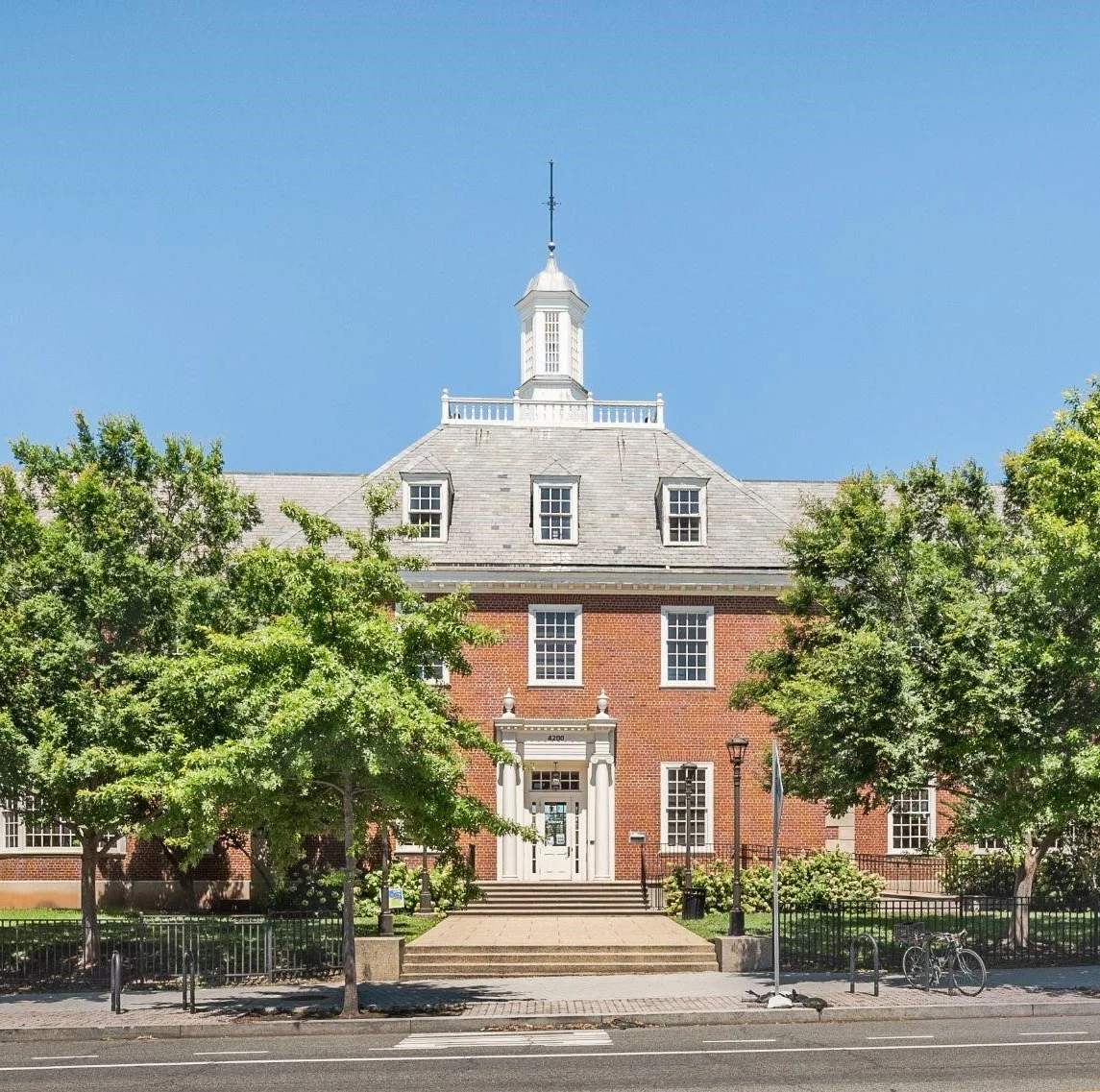 A large brick building with a stepped roof and a white tower on top, surrounded by green trees and a sidewalk, under a clear blue sky.