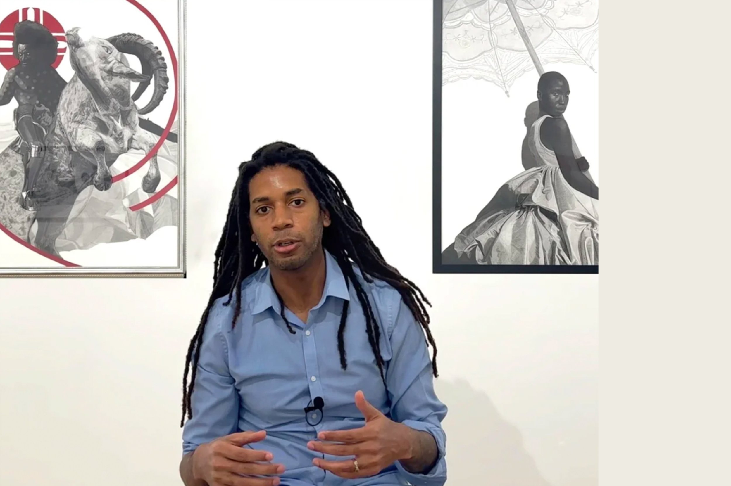 Man with long dreadlocks wearing a blue shirt sitting in front of two black and white artwork posters on a white wall.