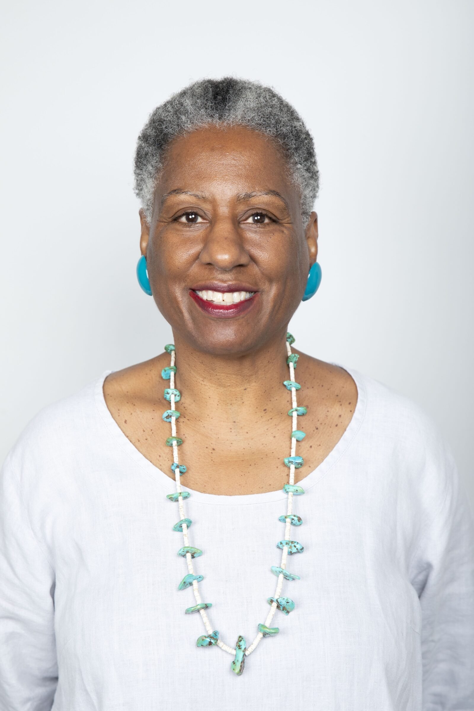 A smiling Black woman with short gray hair, wearing turquoise earrings, a matching turquoise necklace, and a white top, standing against a plain white background.