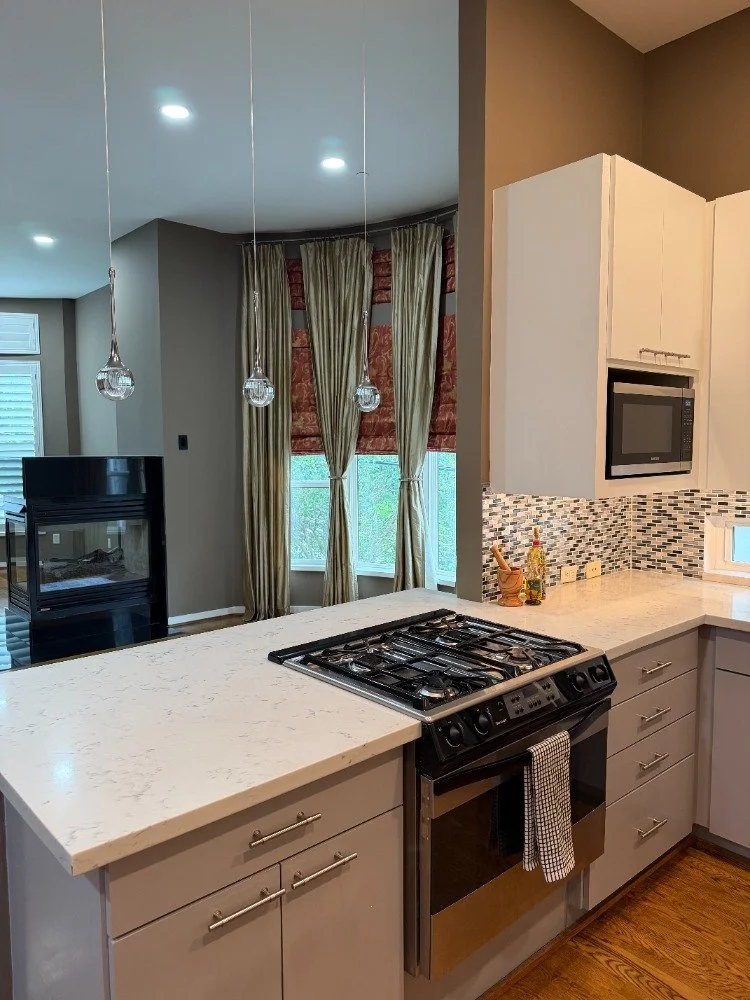 Kitchen with beige cabinets, white marble countertop, stainless steel gas stove, mosaic tile backsplash, and pendant lights hanging from the ceiling. There are curtains and a window in the background with a fireplace and a window with blinds.