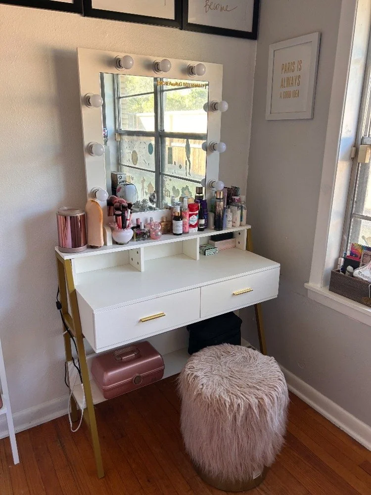 White vanity table with a mirror surrounded by light bulbs, holding various skincare and makeup products. A fluffy pink stool is in front, and a pink makeup case is on the floor below. A framed quote on the wall reads 'Paris is always a good idea,' and a window nearby shows a sunny outdoor scene.