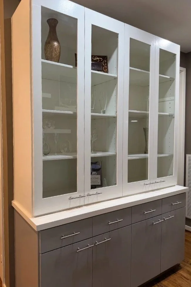 A white and gray china cabinet with glass doors, displaying decorative glassware and vases inside, situated in a room with wooden flooring.