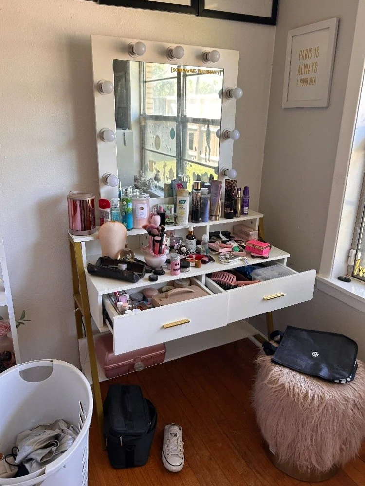 A white makeup vanity table with a mirror surrounded by light bulbs, cluttered with various cosmetics and beauty products. There is a large framed quote on the wall to the right, and a window with a view outside. Items like a pink makeup organizer, brushes, perfume bottles, and makeup palettes are visible. A white laundry basket and a fluffy pink stool with a black bag and a laptop on top are in front of the table.
