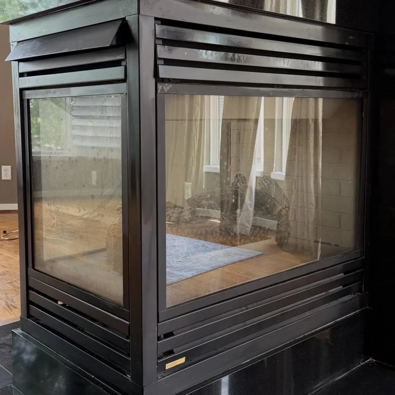 Black metal fireplace with glass panels in a living room, reflecting a sofa, curtains, and windows.