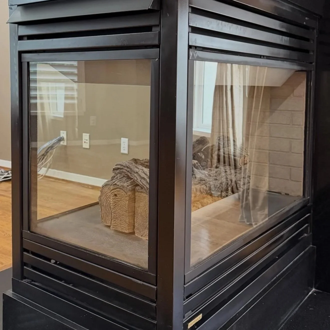 Close-up of a modern black metal and glass fireplace with a log inside, in a cozy living room with curtains and a window reflected in the glass.