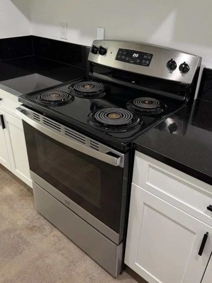 A kitchen stove with four coil burners, a digital control panel, and a stainless steel oven door, surrounded by white cabinets and black countertops.