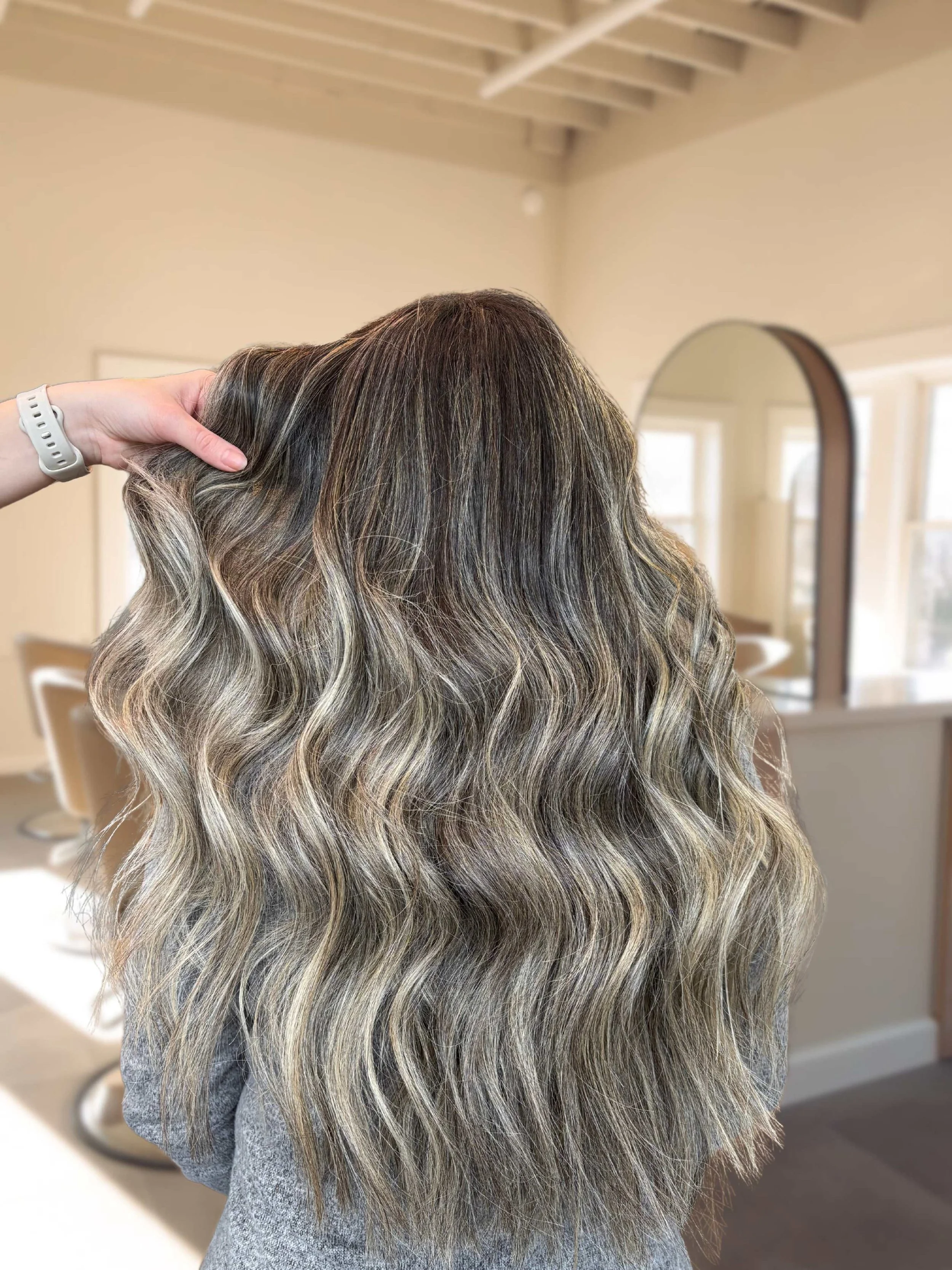 A woman with long wavy hair featuring dark roots and blonde highlights, held by a hand inside a bright, modern salon.