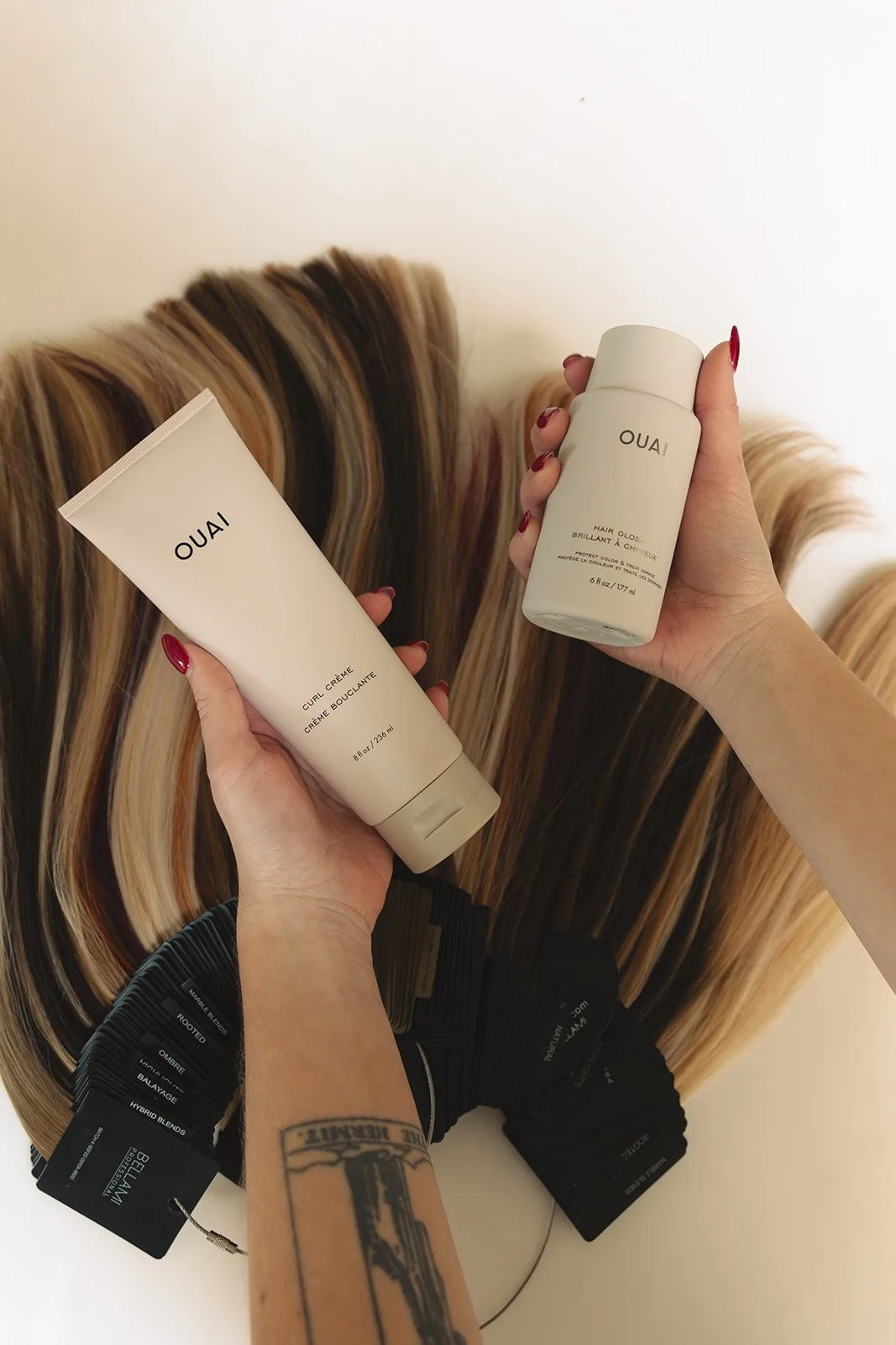 Hands holding two OUAI hair products in front of a wall of hair color swatches and hair color samples.
