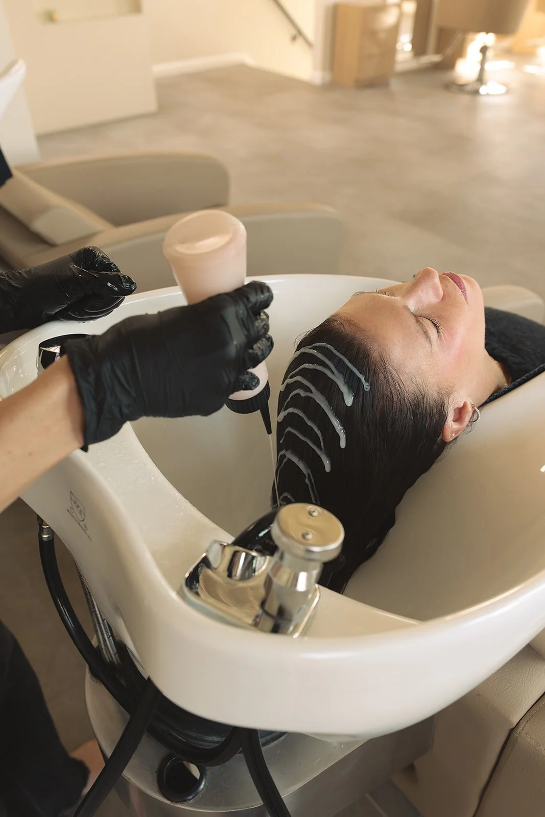 A woman reclines with her head in a salon sink while a hairstylist applies hair treatment or dye, wearing black gloves and holding a bowl or container.