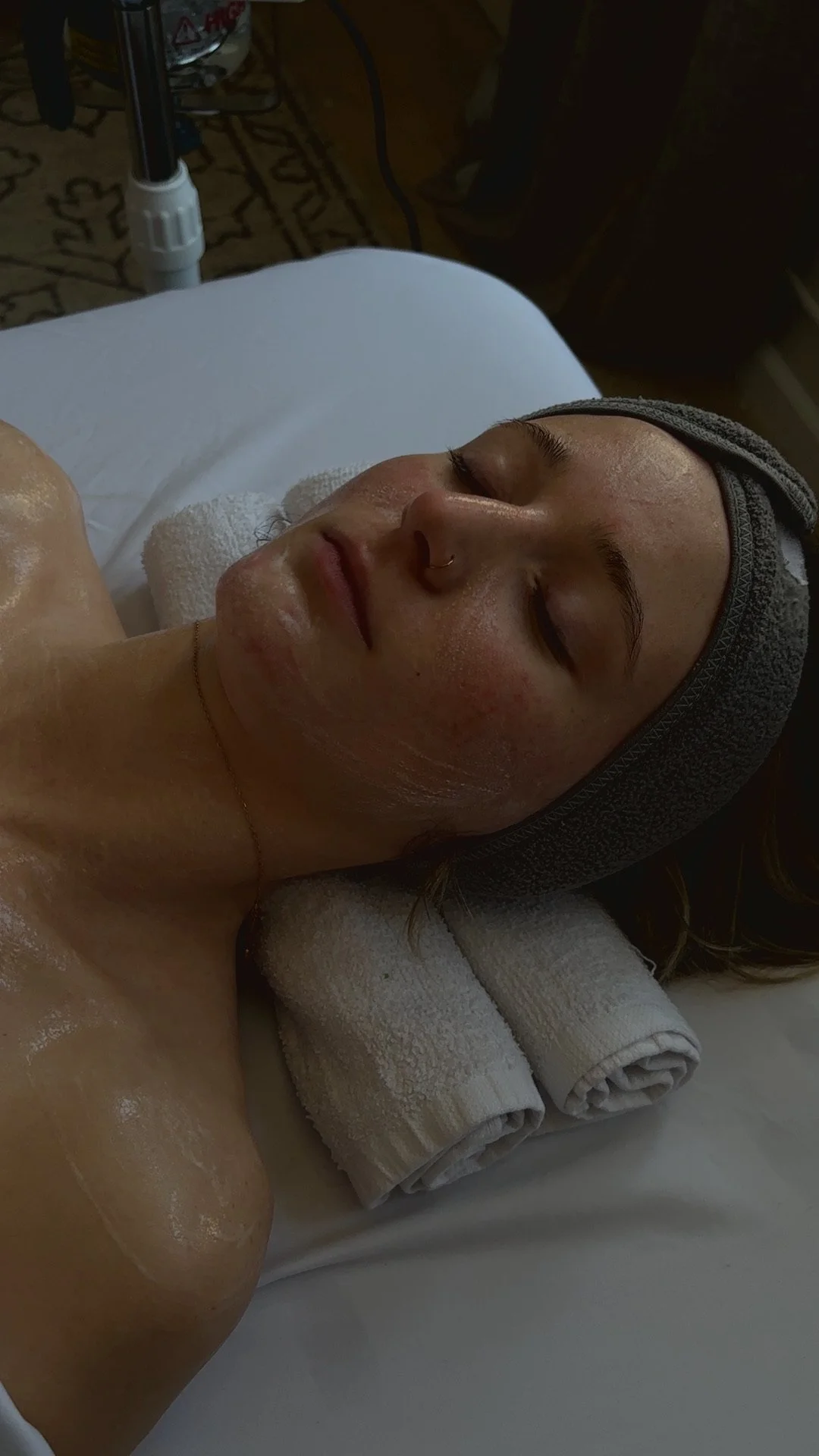 A woman lying on a treatment bed with a towel under her head, wearing a gray headband, with her eyes closed, receiving a relaxing glow facial treatment.