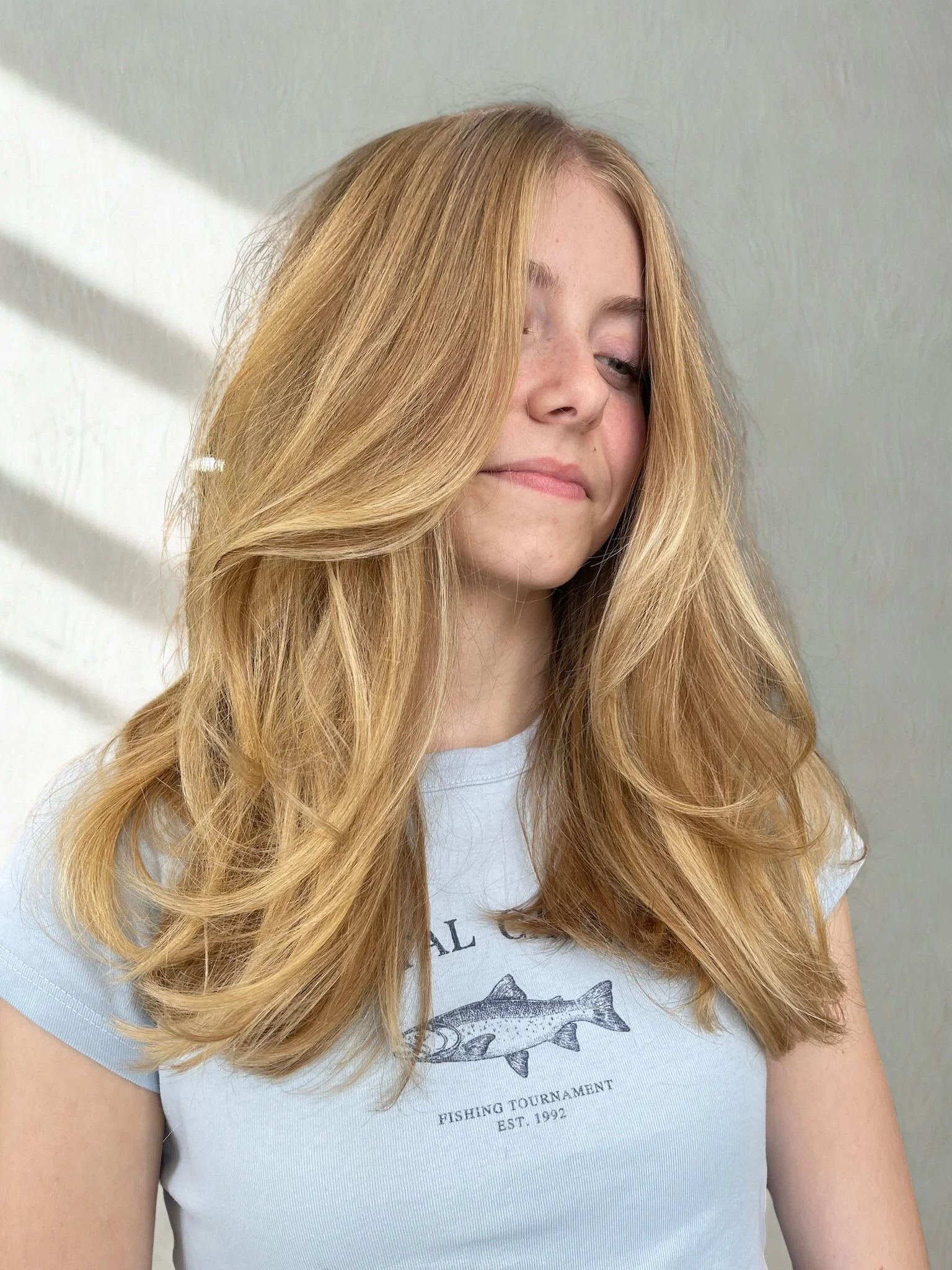 A young woman with long, wavy blonde after a blowout hair partially covering her face, standing against a light-colored wall with soft shadows.