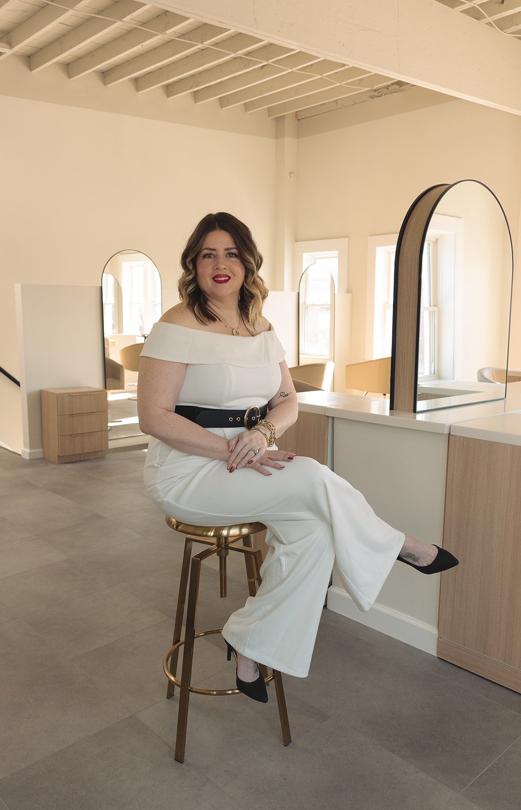 Janelle, Bobbi's staff aesthetician, with shoulder-length wavy hair, wearing a white off-shoulder jumpsuit with wide-legged pants and black high heels, sitting on a gold stool in a bright, modern room with multiple mirrors and windows.