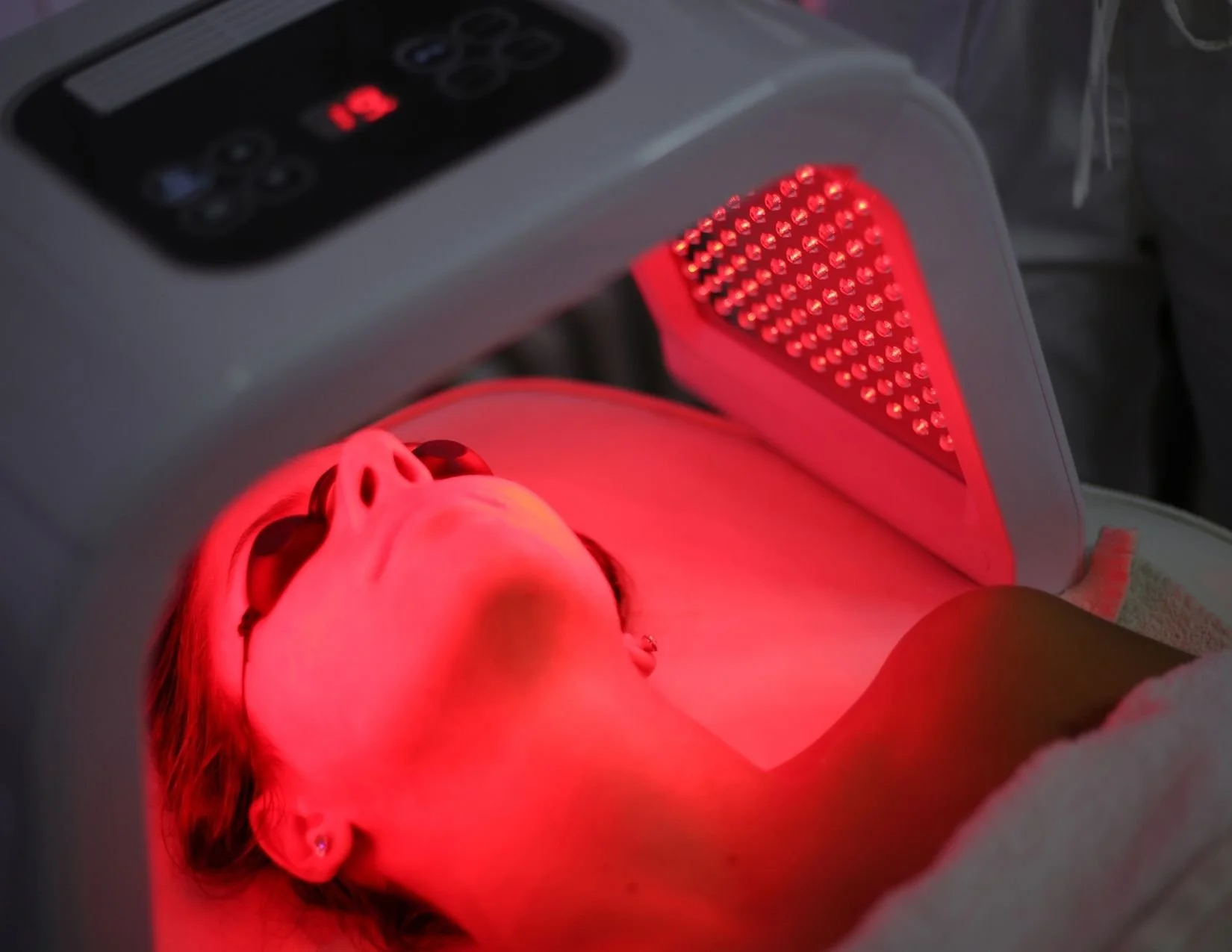 A woman lying under a red LED light therapy device on her neck, wearing sunglasses during a facial treatment.