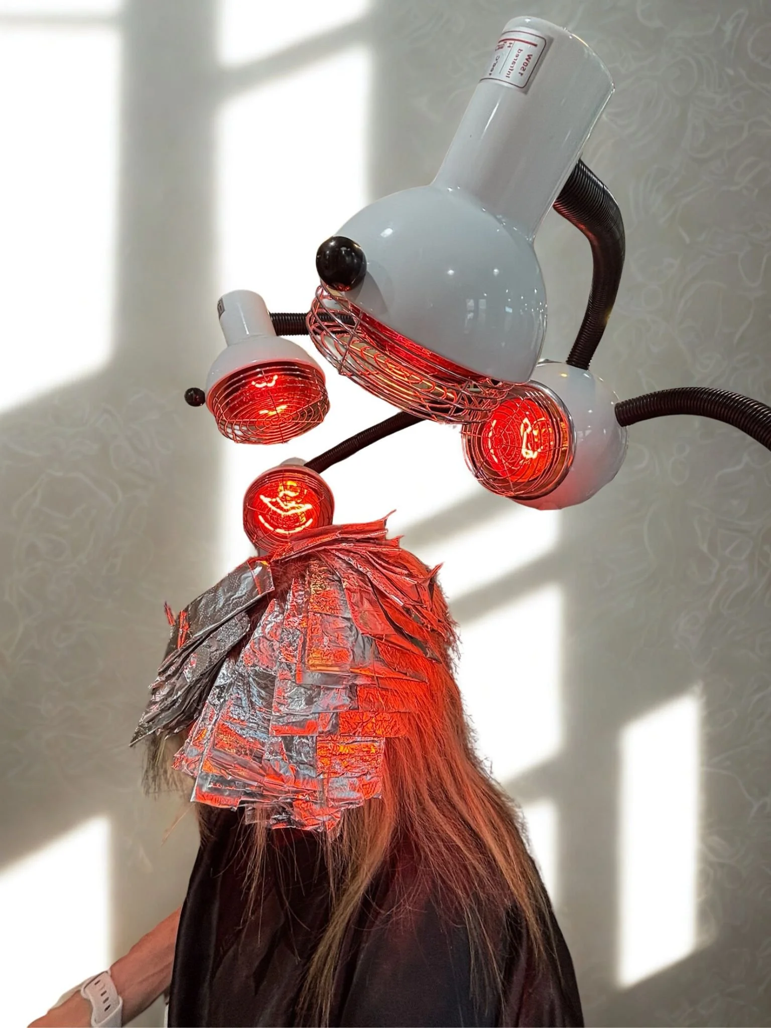 Person receiving a hair treatment with a machine emitting red heat light, with a metallic foil covering part of their hair, in a professional setting.