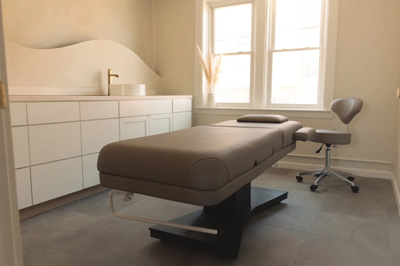A massage table in a treatment room with natural light from a window, white cabinetry, a vase with pampas grass, and a modern swivel chair.