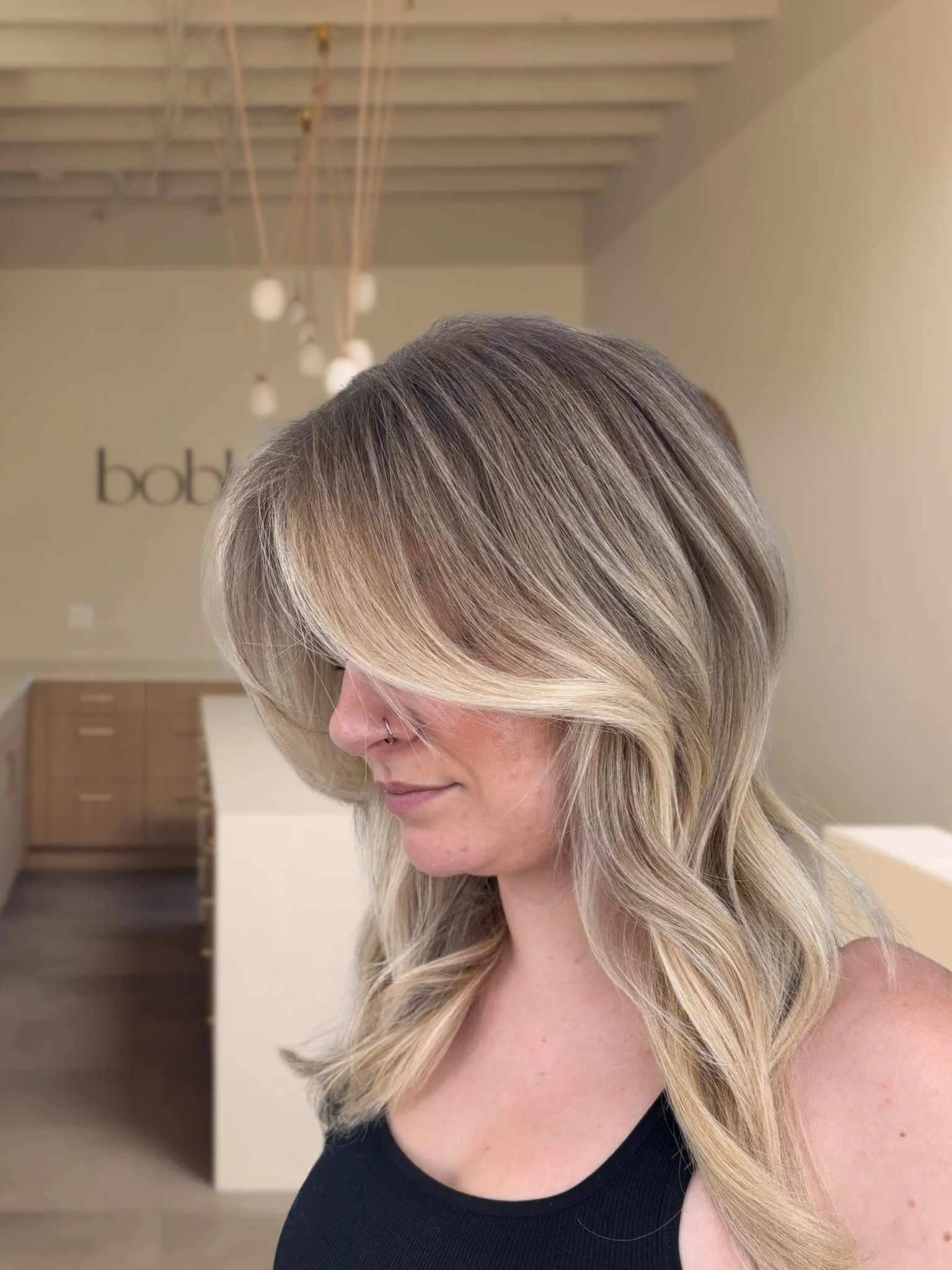 Side profile of a woman with blonde, layered, wavy hair and a nose piercing, inside a modern salon with wooden drawers and hanging light fixtures.