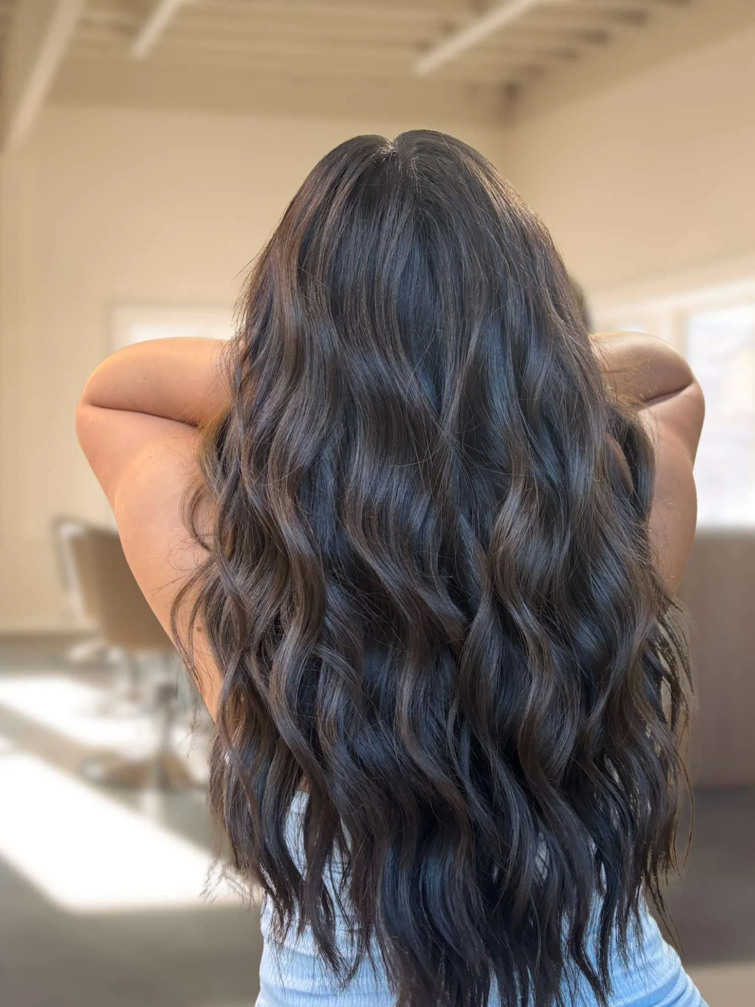 Back view of a person with long, wavy dark brown hair in a bright room with beige walls and windows.