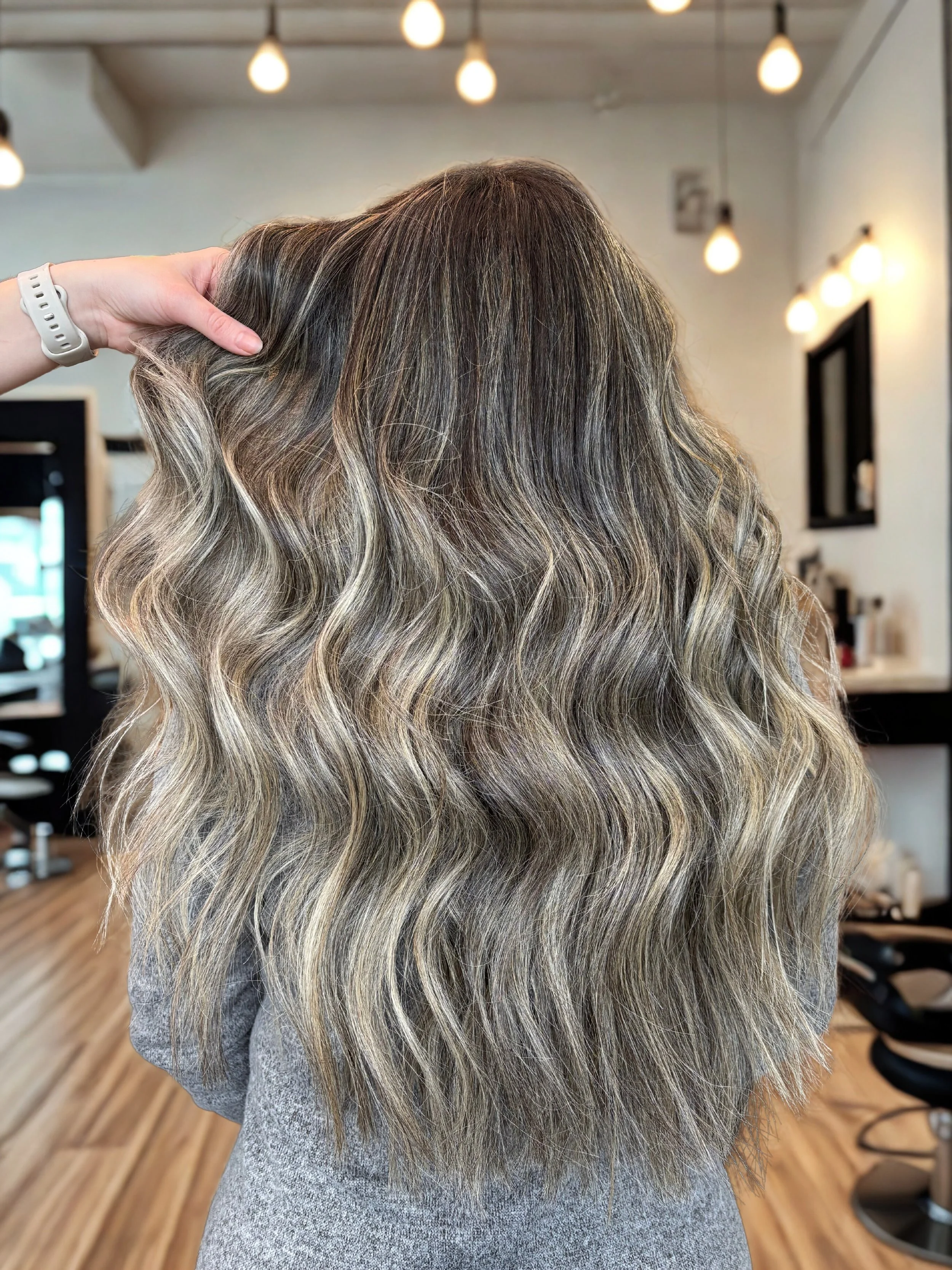 Back view of a woman with long, wavy, blonde-brown hair with beautiful balayage highlights in a salon setting.