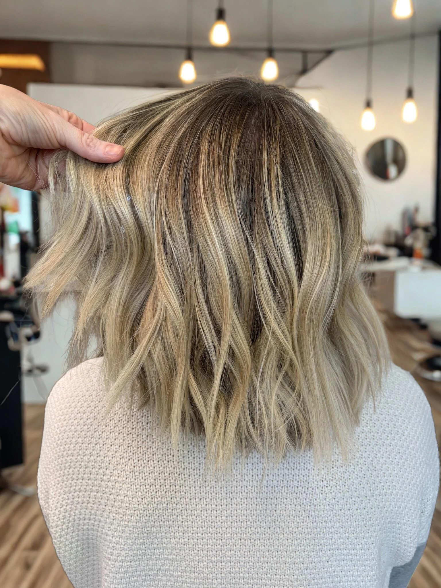 Back of a woman's head with shoulder-length blonde, wavy hair in a salon, held by a stylist's hand, with salon lighting and decor in the background.