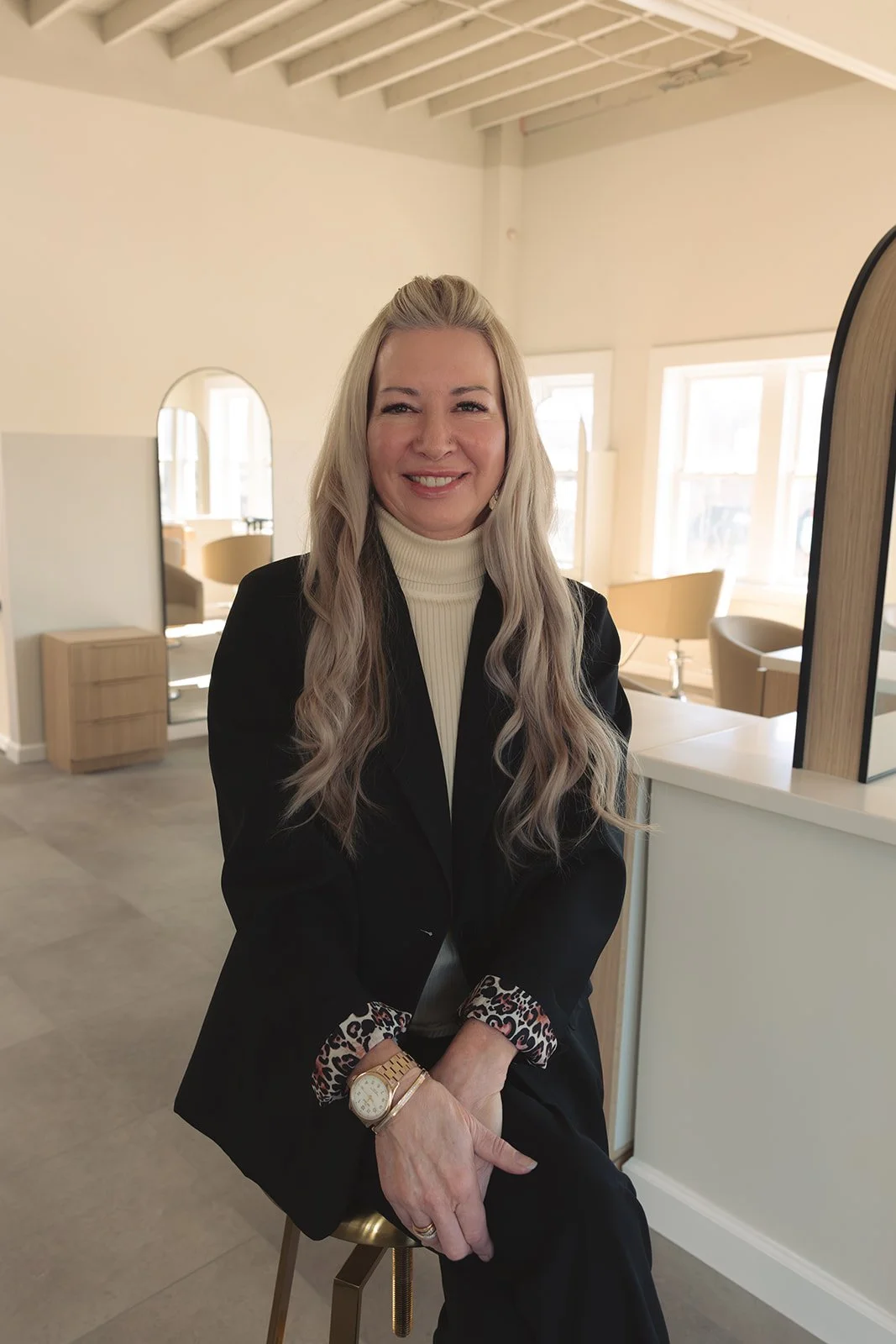 Rebecca, Bobbi's staff RN, with long, wavy blonde hair, wearing a black blazer with leopard print cuffs, a cream turtleneck, and a watch, sitting on a stool in a bright, modern indoor space with large windows and minimalist decor.