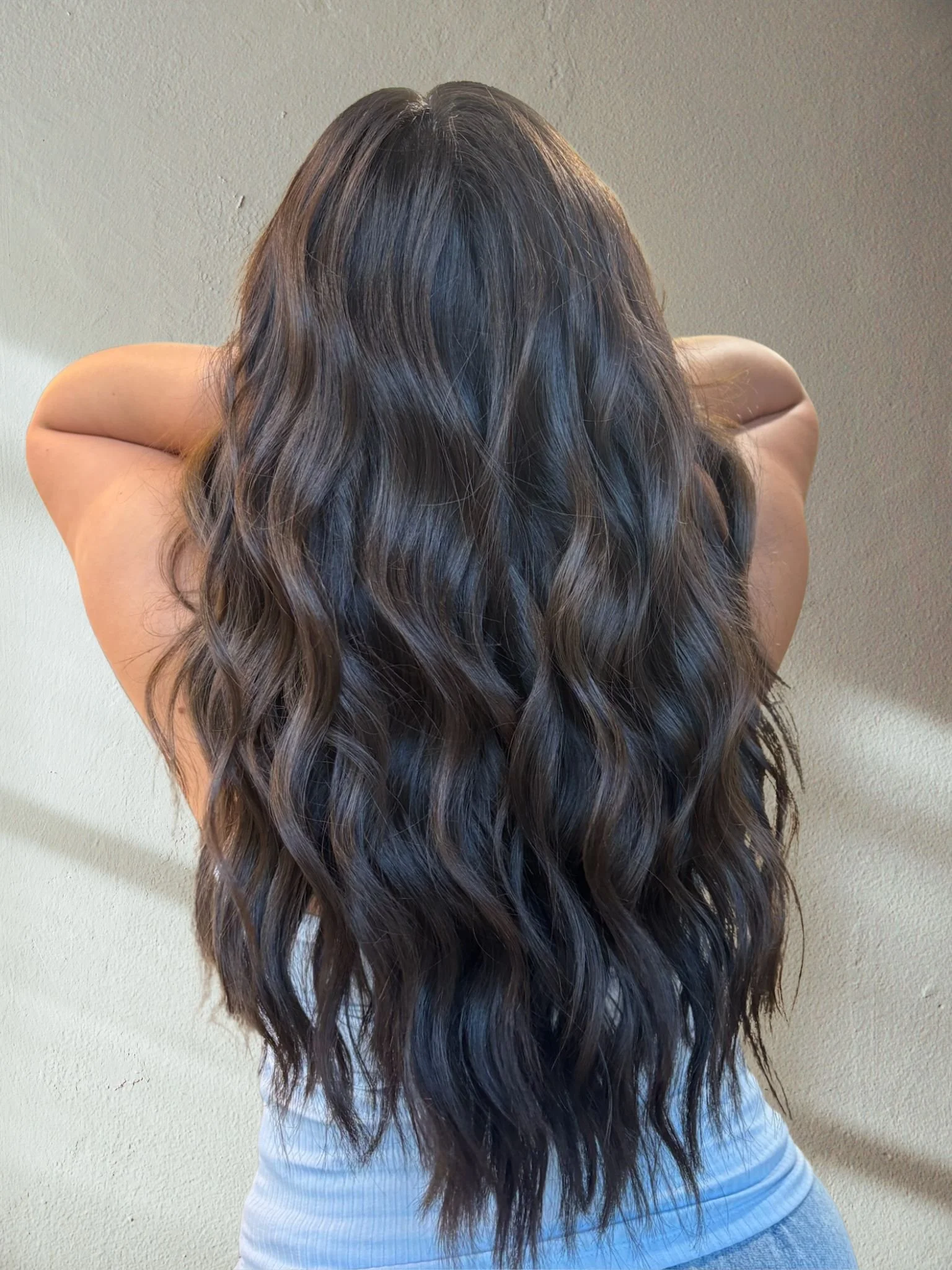A woman with long, wavy dark brown custom hair color , standing with her back to the camera against a plain wall.