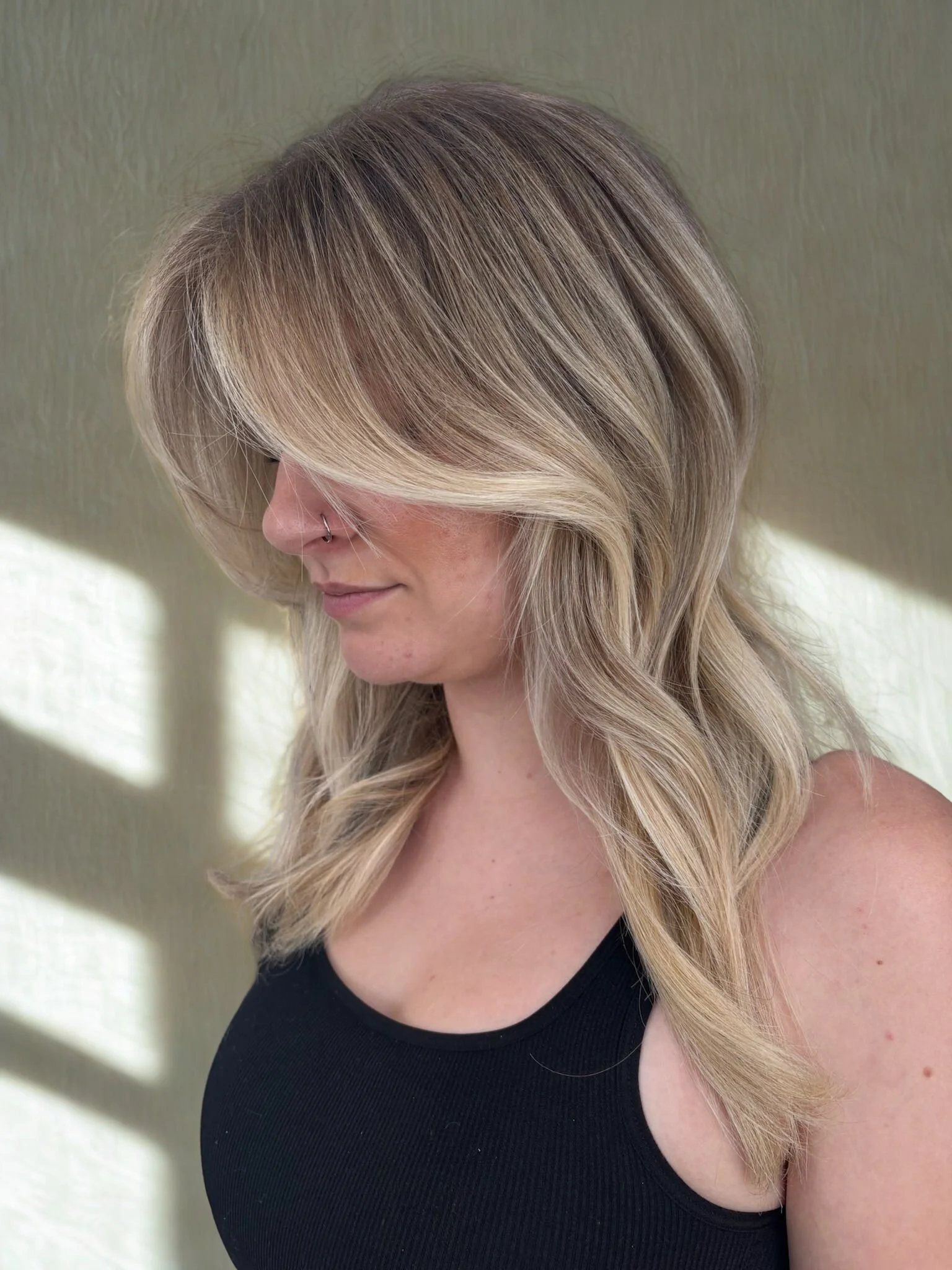 Side profile of a woman with custom blonde balayage layered hair, wearing a black sleeveless top, standing in front of a textured wall.
