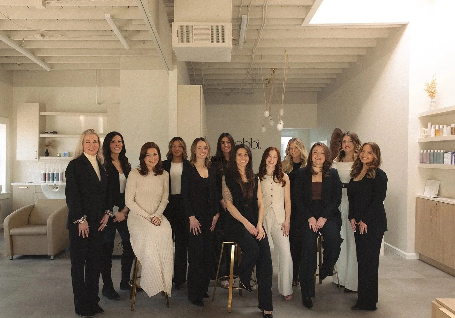 Bobbi staff, Grosse Pointe Premier Salon, in an indoor space, smiling at the camera. The room has a modern, minimalist design with neutral colors, shelves with bottles and jars, and hanging pendant lights.