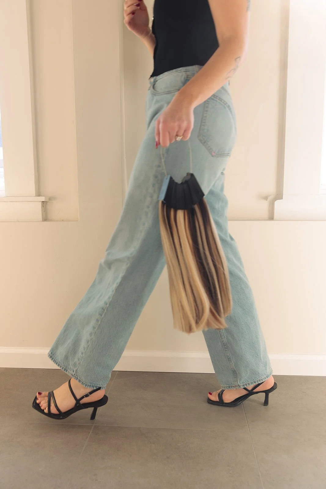A person wearing black strappy high heels, loose light-wash jeans, and a black top holding custom hair extensions.