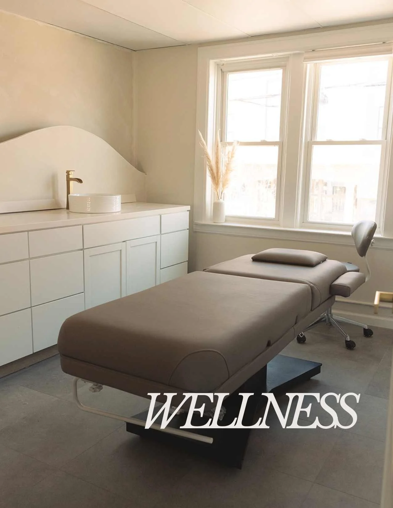 A massage or therapy table in a bright, minimalist room with large windows, white cabinetry, and decorative dried grasses in a vase. The word 'WELLNESS' is overlaid at the bottom of the image.